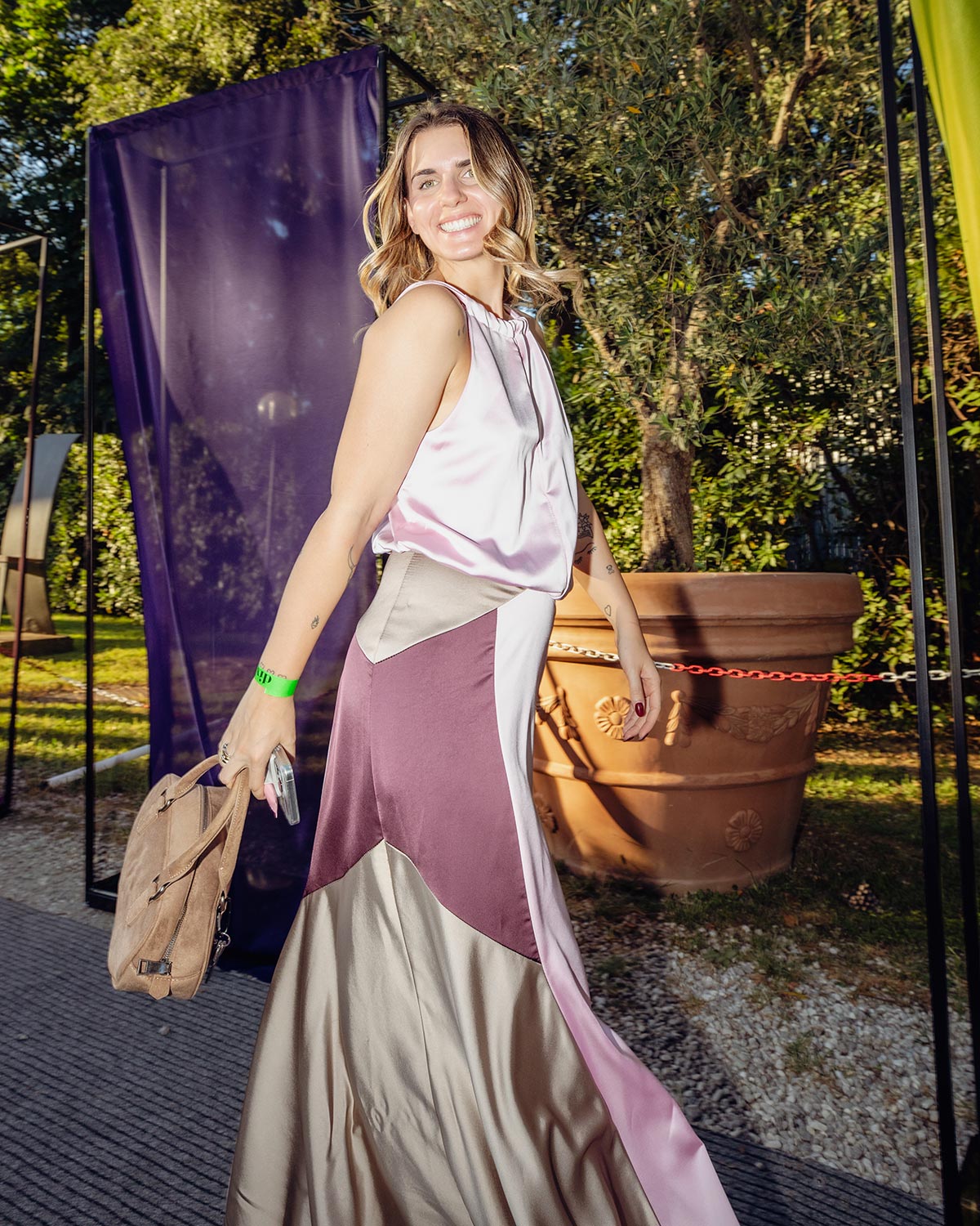 Smiling woman in a colour-block satin evening dress holding a tan bag at an outdoor garden event
