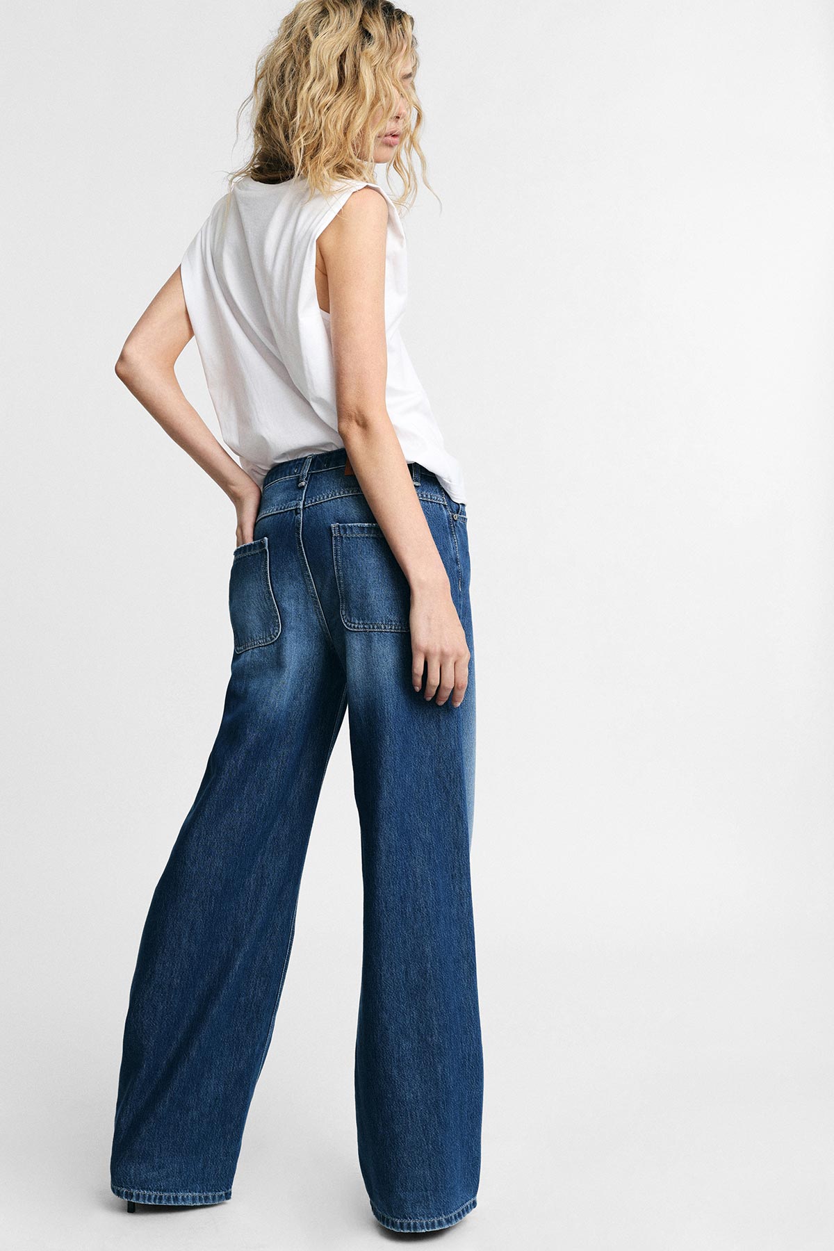 Woman in white sleeveless top and high-waisted wide-leg blue jeans, back turned and looking over shoulder against plain studio background