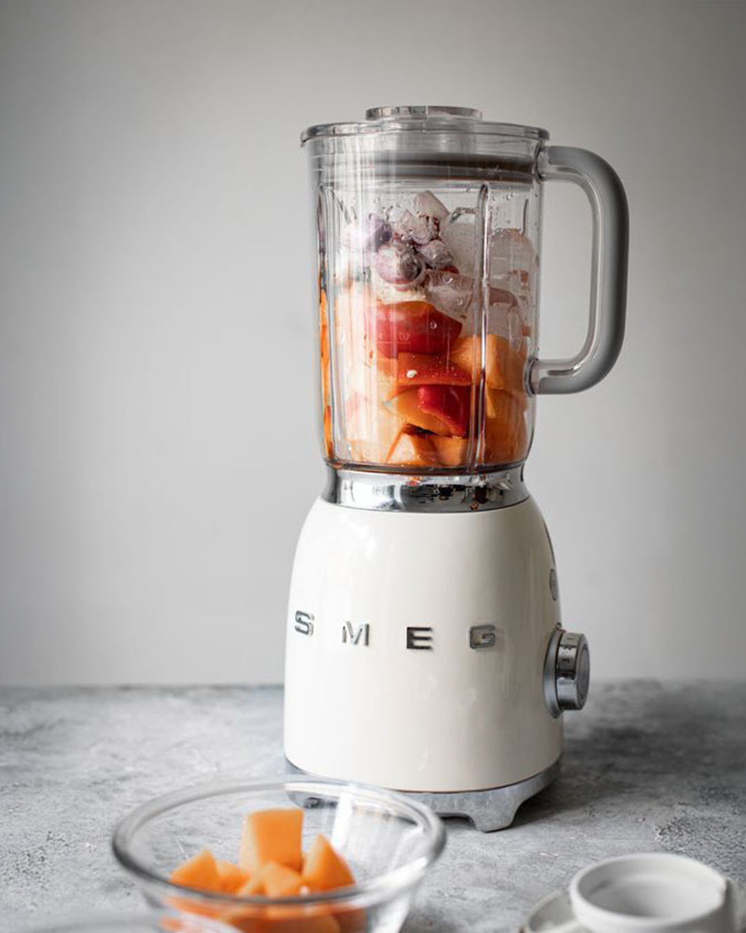 Cream SMEG countertop blender filled with chopped orange fruit, grapes and ice on a grey surface; clear glass bowl with melon cubes in the foreground