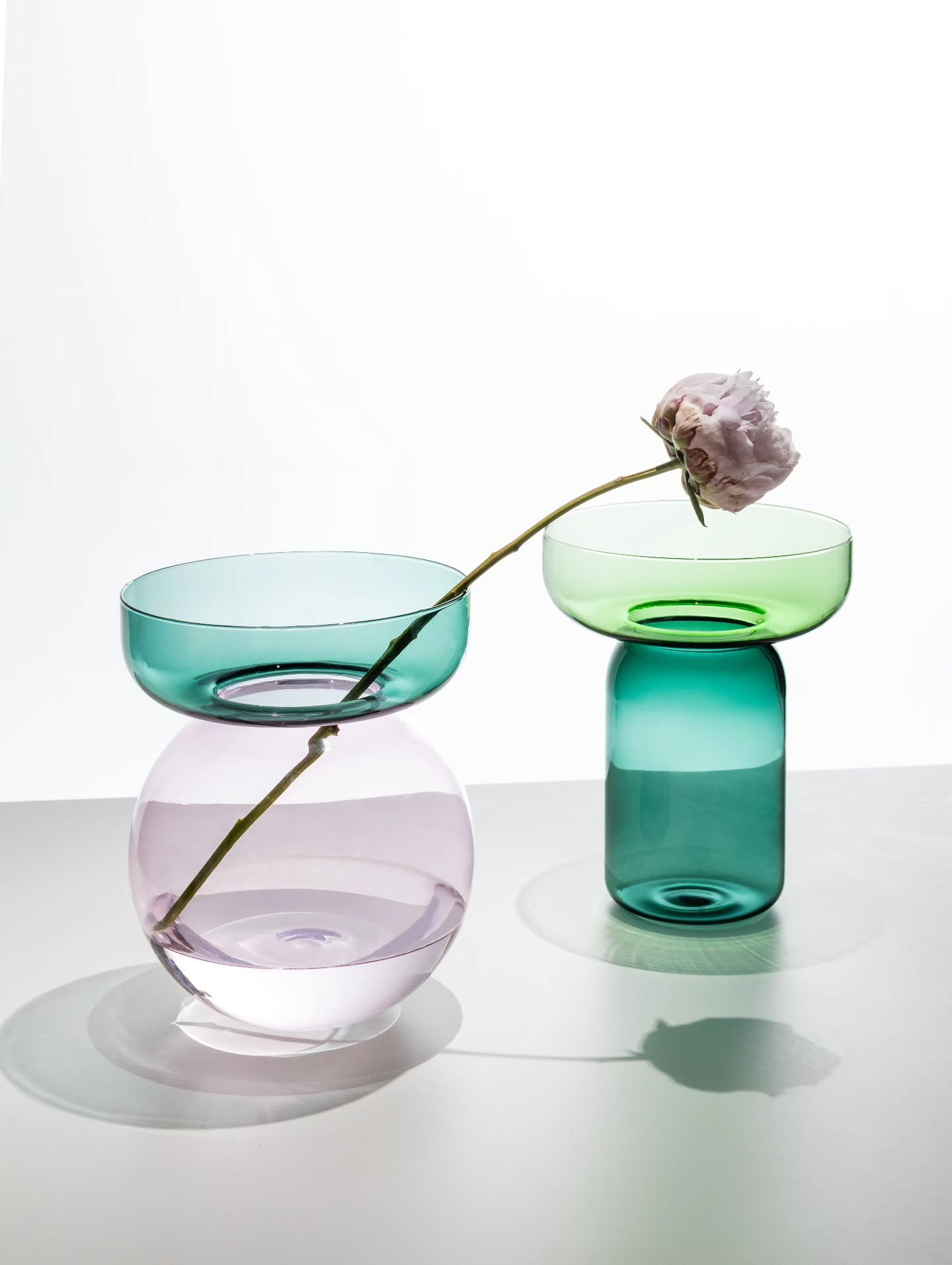 Two modern coloured glass vases (teal and green) with a single pale pink peony draped across them on a white background