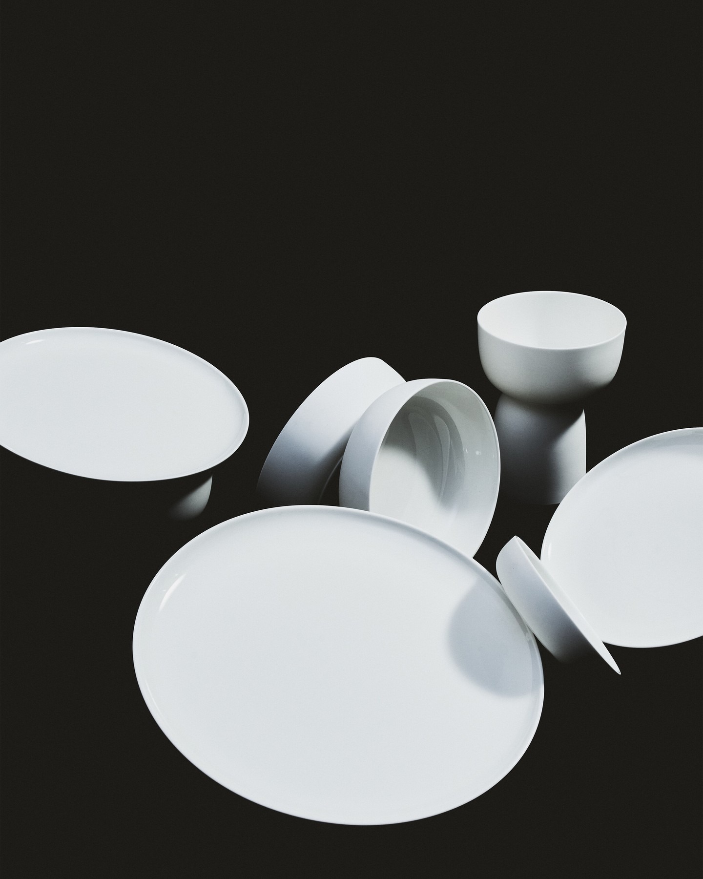 Minimalist white ceramic plates and bowls scattered and stacked on a black background, studio-lit modern tableware