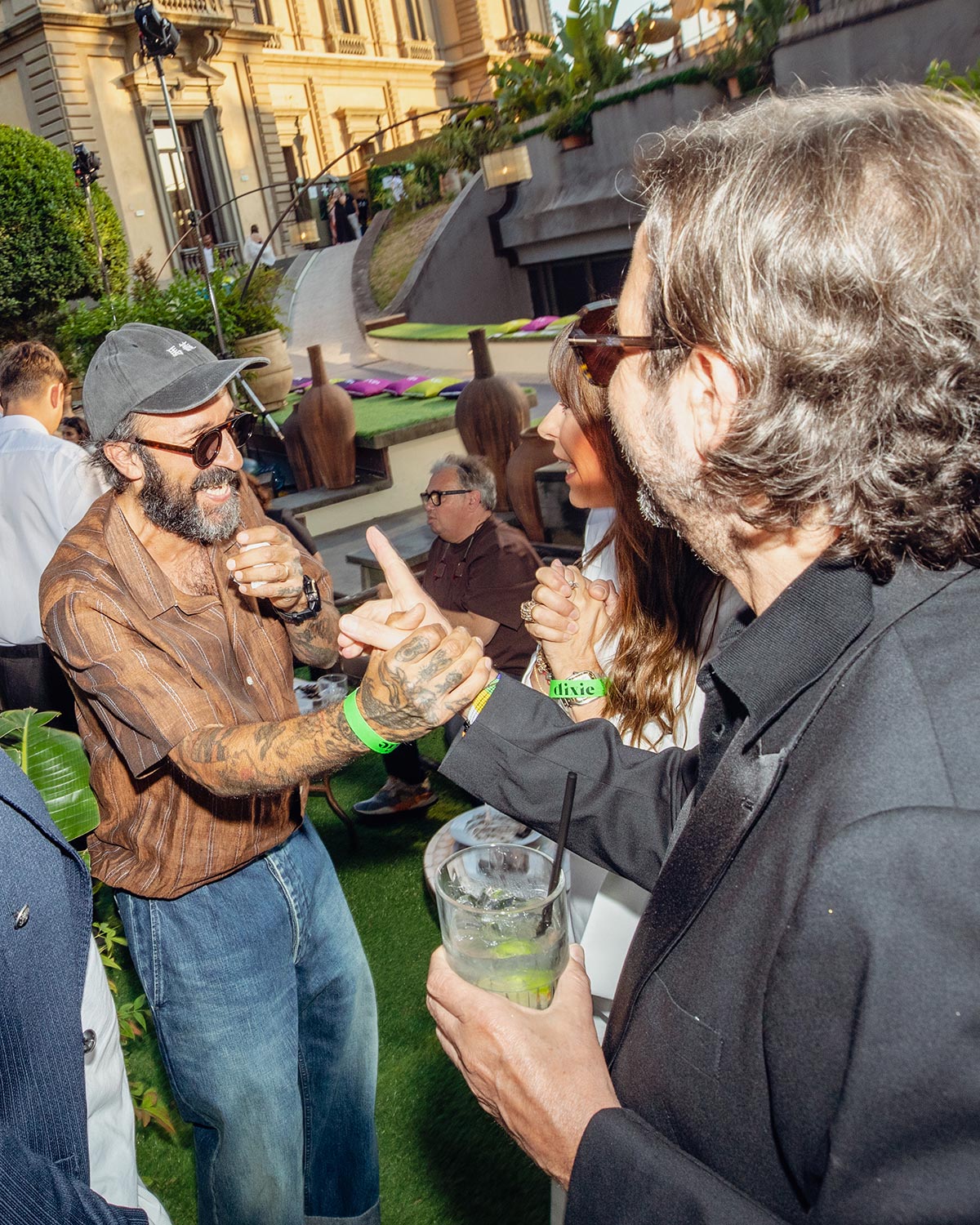 People greeting and shaking hands at a rooftop garden party, tattooed man in cap, sunglasses, cocktail
