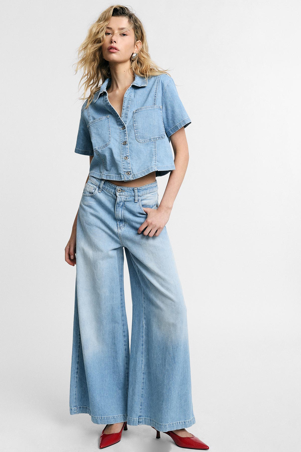 Model in light blue cropped denim shirt and high-waisted wide-leg jeans, posing against white background wearing red heeled shoes