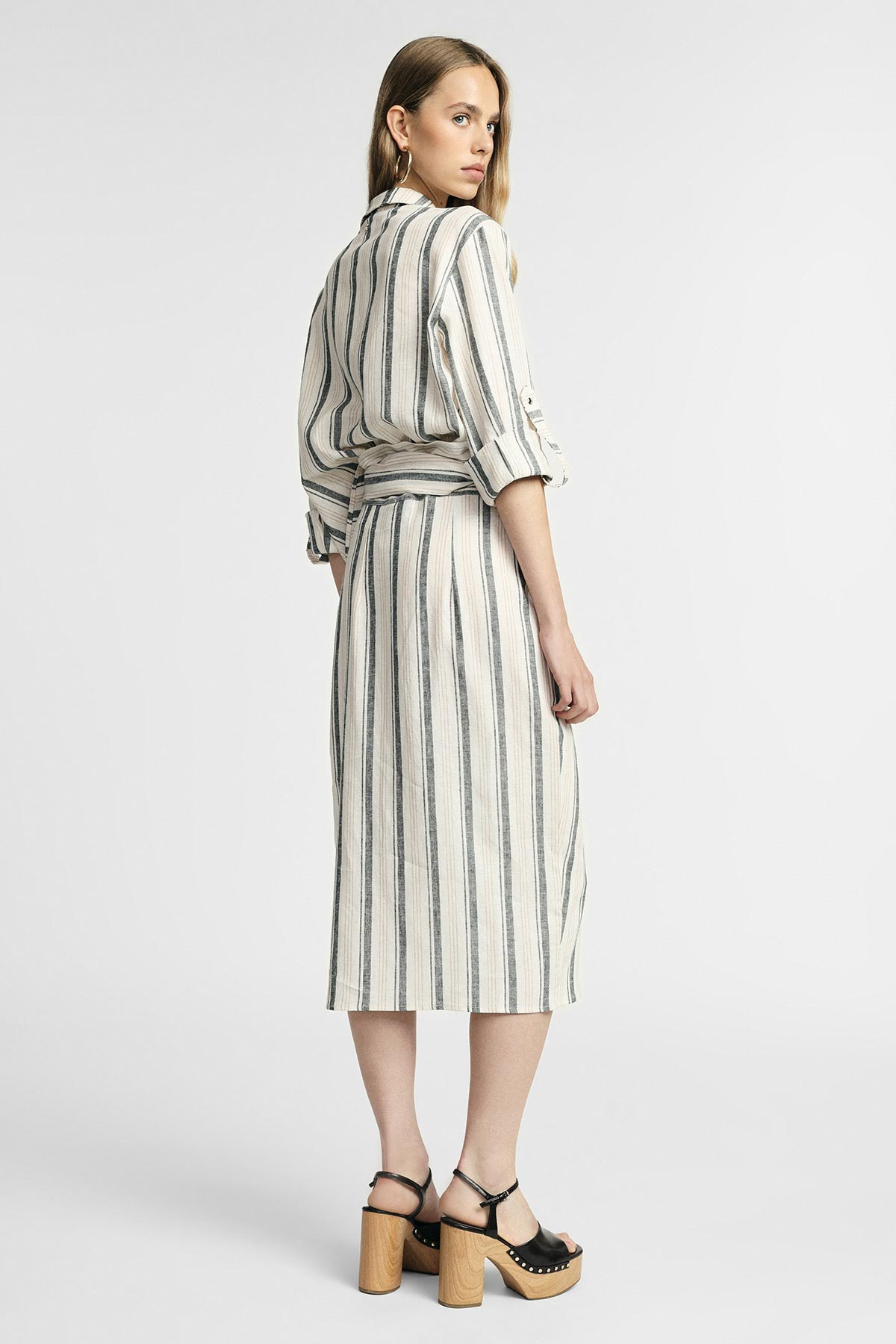 Back view of model wearing cream and navy vertical-striped midi shirt dress with tied waist, looking over shoulder, wooden platform sandals