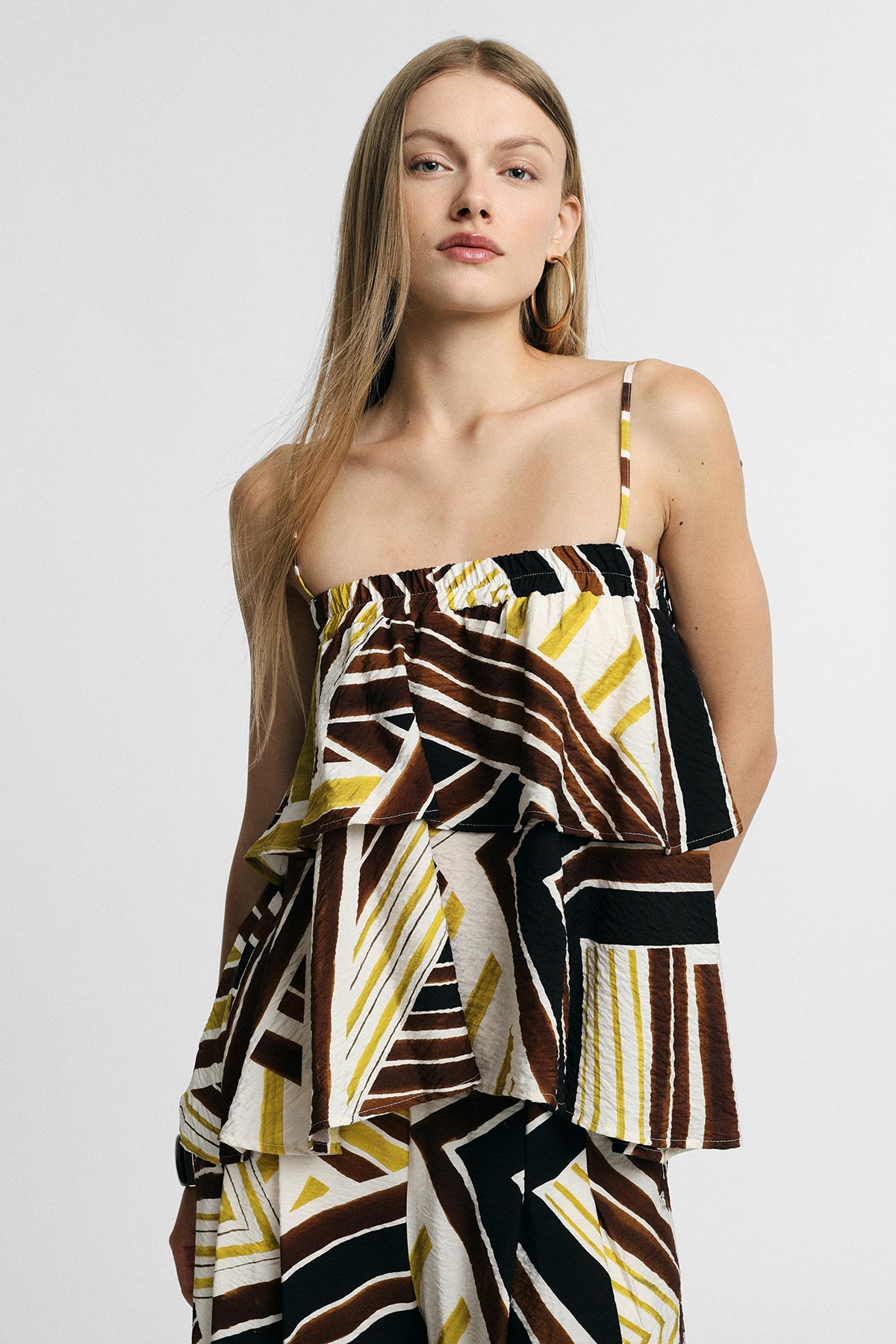 Woman in abstract brown, black and yellow patterned tiered summer dress with thin straps and hoop earring on plain background