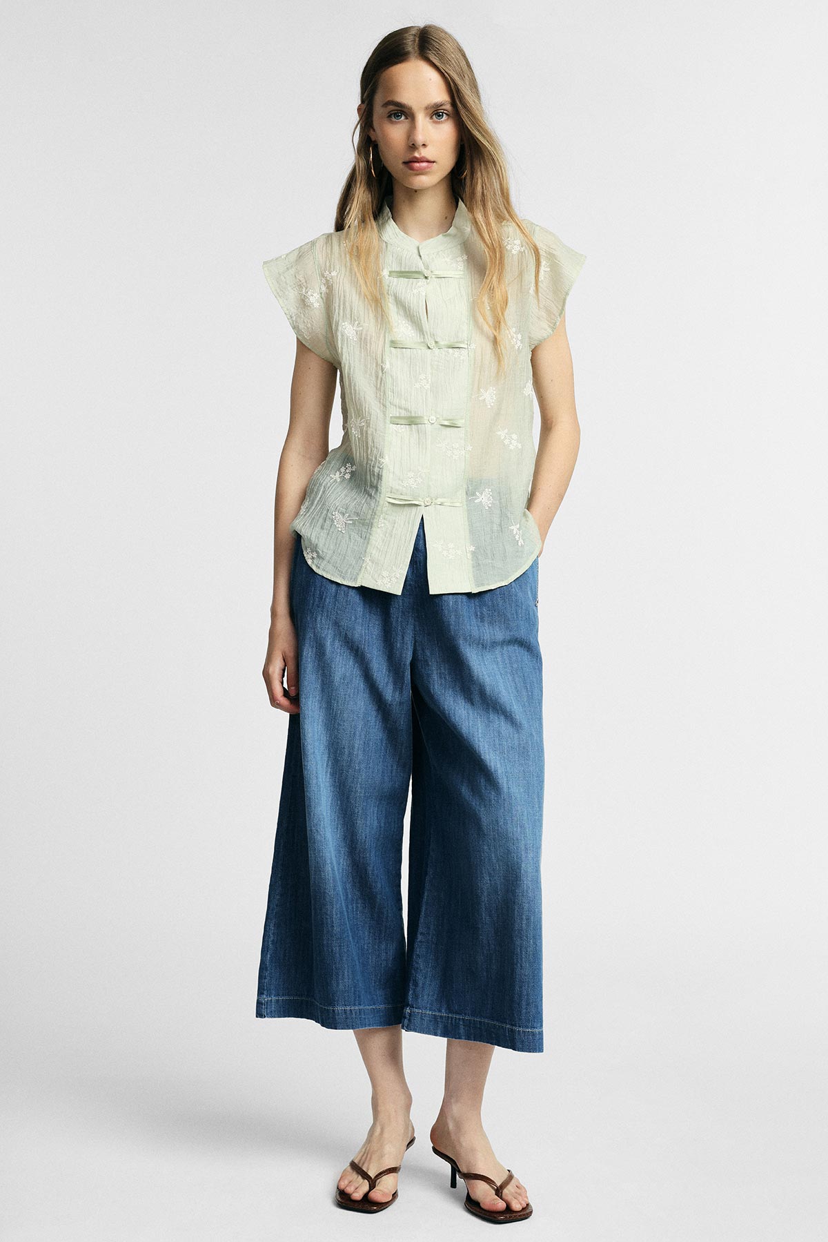 Female model wearing a light green sheer Mandarin-collar blouse with frog closures, wide-leg cropped denim trousers and black sandals