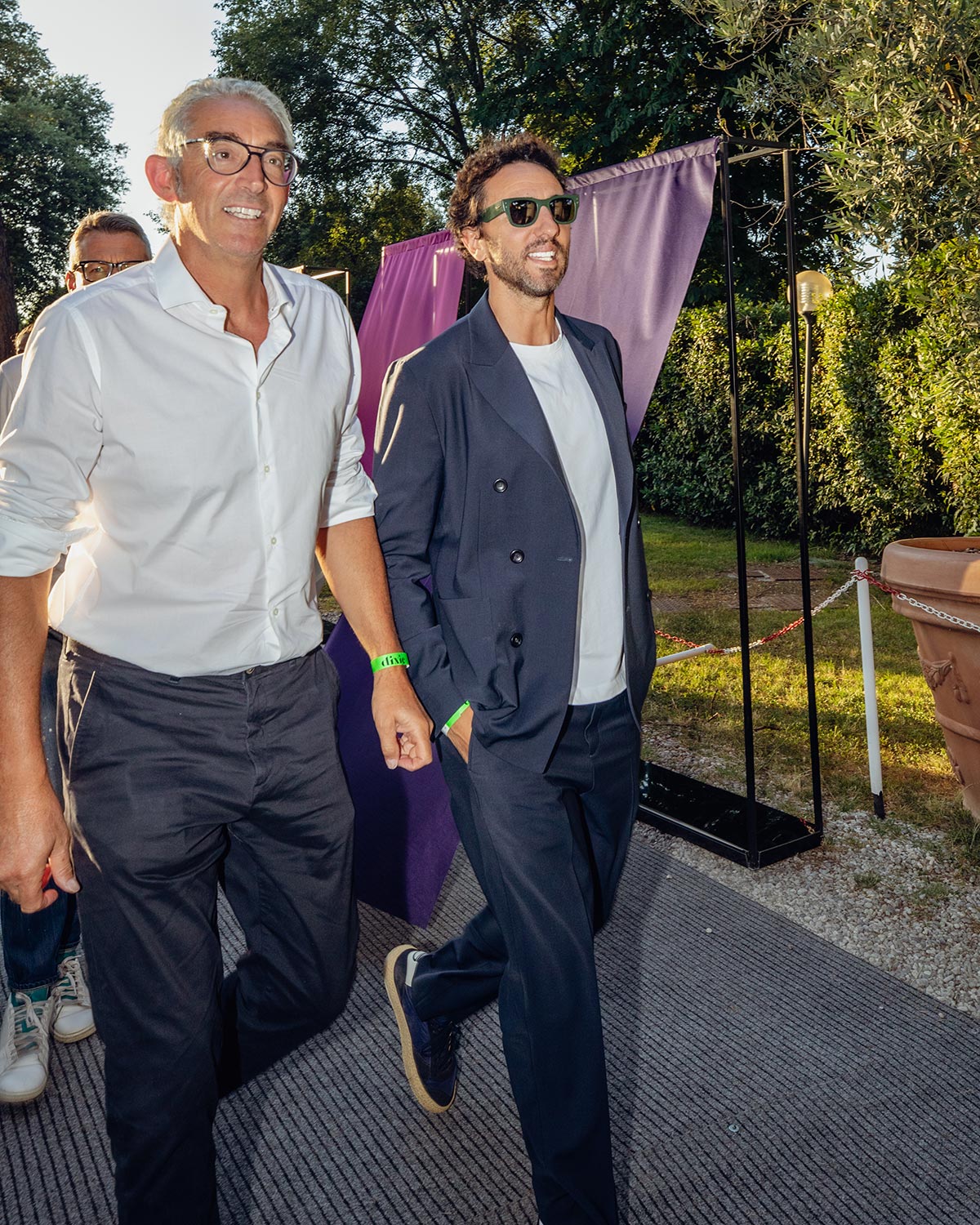 Two smiling men walking at an outdoor event by a purple backdrop and trees; one in a white shirt, the other in a navy suit and sunglasses