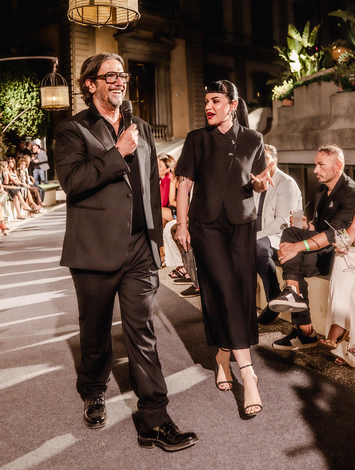 Smiling man with microphone and female co-host in black walking runway past seated guests during evening fashion show