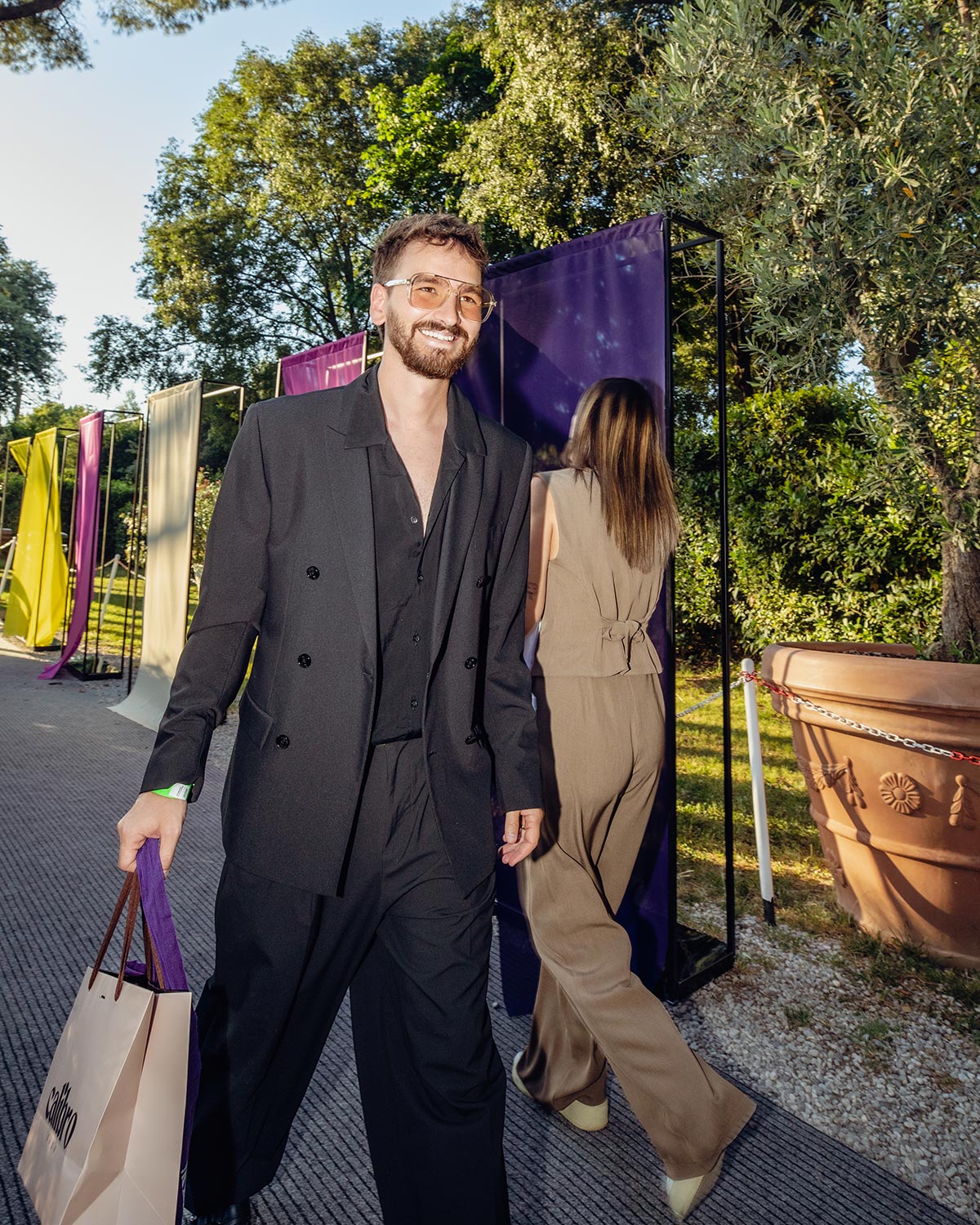 Smiling man in black double-breasted suit carrying a shopping bag at an outdoor fashion event with colourful fabric panels