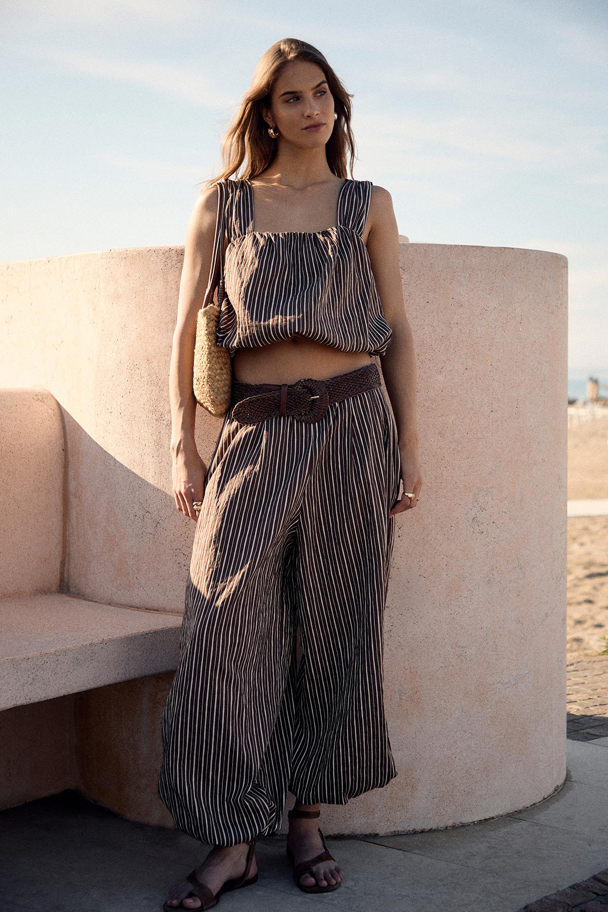 Woman standing by curved wall at beach wearing striped crop top and wide-leg trousers, straw shoulder bag