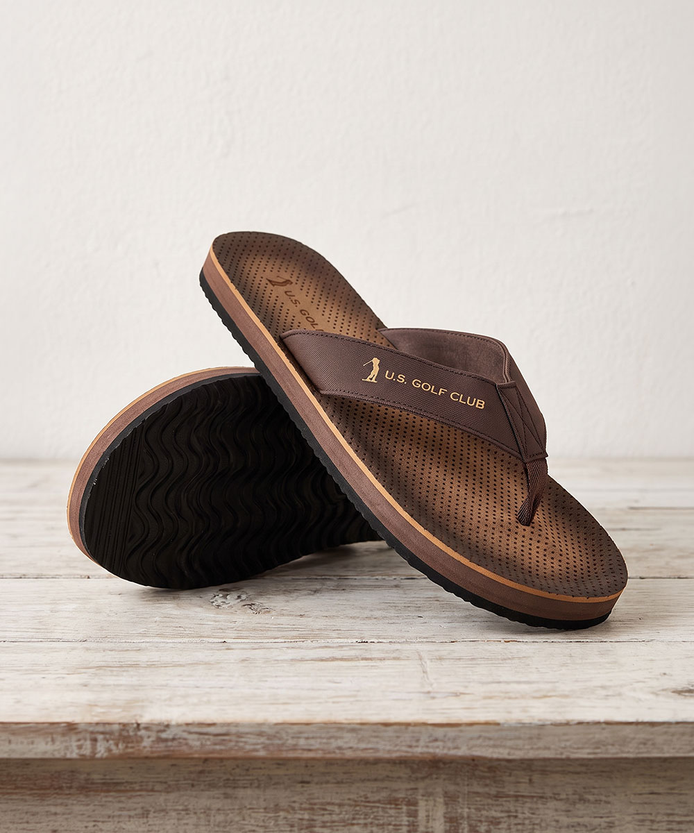 Brown perforated leather flip-flops with U.S. Golf Club logo on strap, cushioned two-tone sole on wooden surface