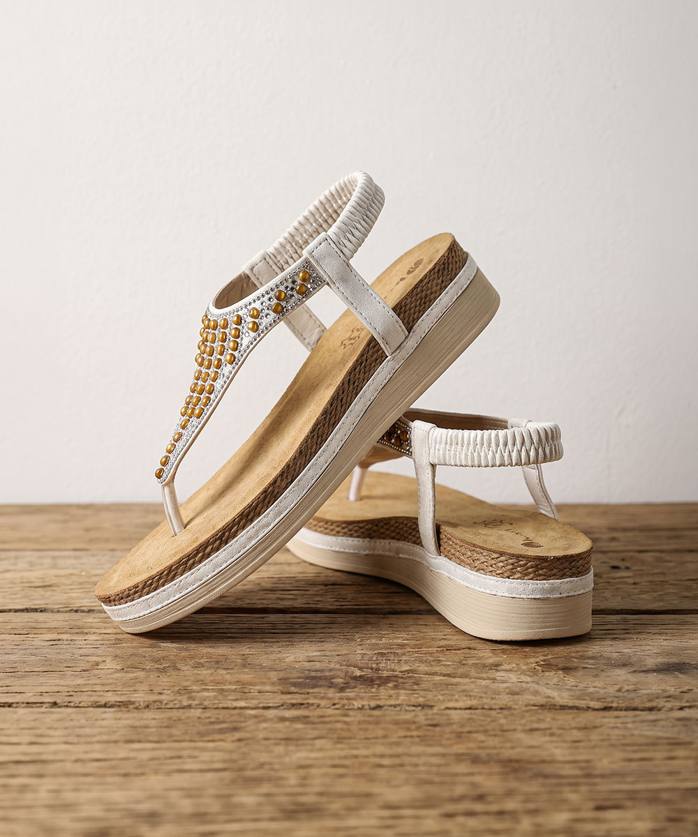 Pair of white women's platform thong sandals with gold bead embellishments and elastic ankle straps on a wooden surface