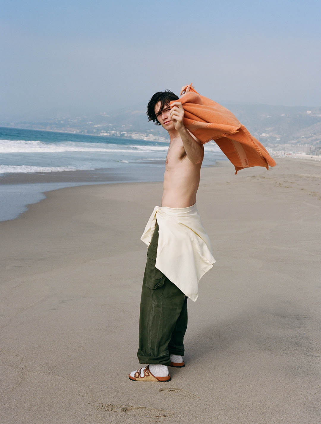 Young man on an empty sandy beach holding an orange towel over his shoulder, wearing green trousers and sandals