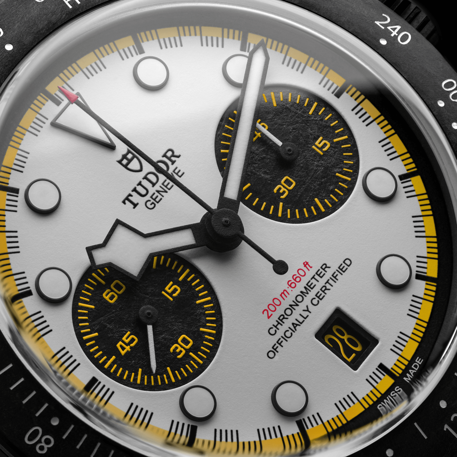 Close-up of Tudor chronograph watch face, white dial with black subdials, yellow accents, date 28 and tachymeter bezel