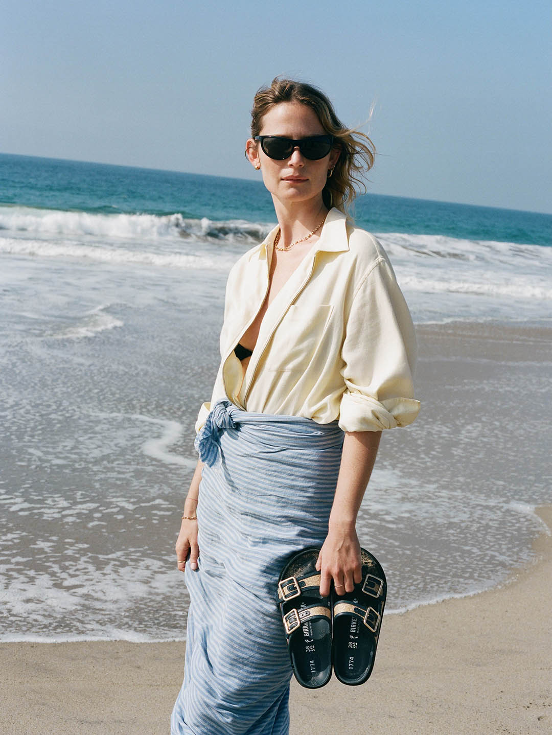 Woman in sunglasses and cream shirt with blue sarong holding black sandals on a sandy beach with ocean waves