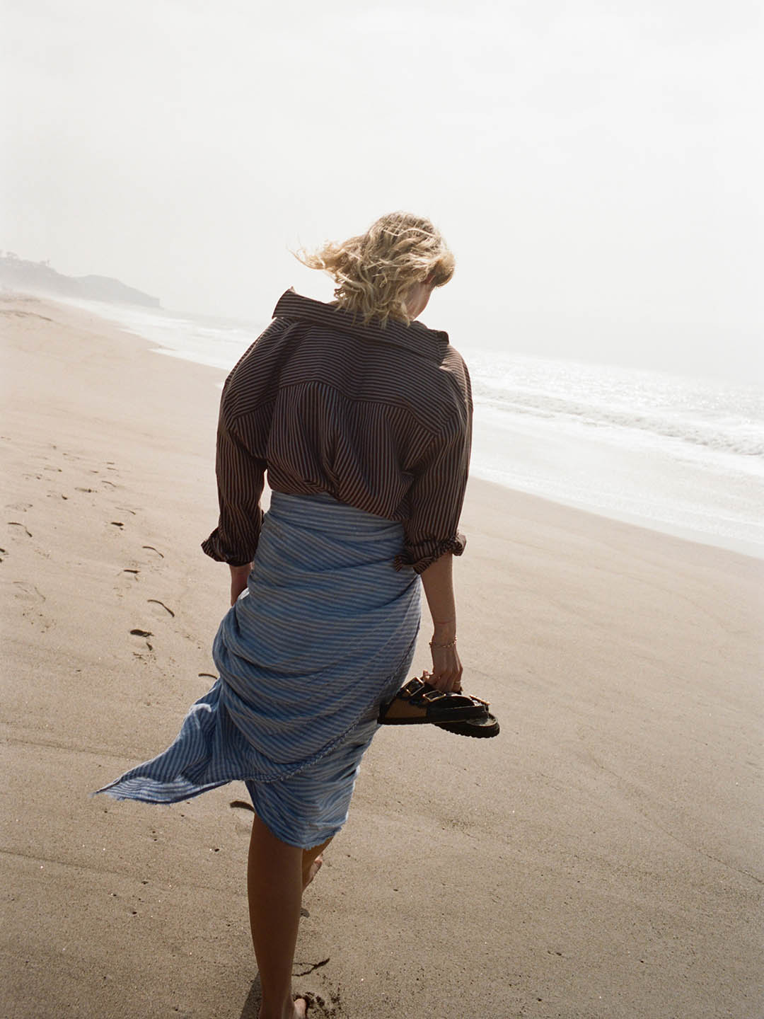 Person walking barefoot along a misty sandy beach, holding sandals and wearing a striped shirt and blue skirt, footprints trailing behind