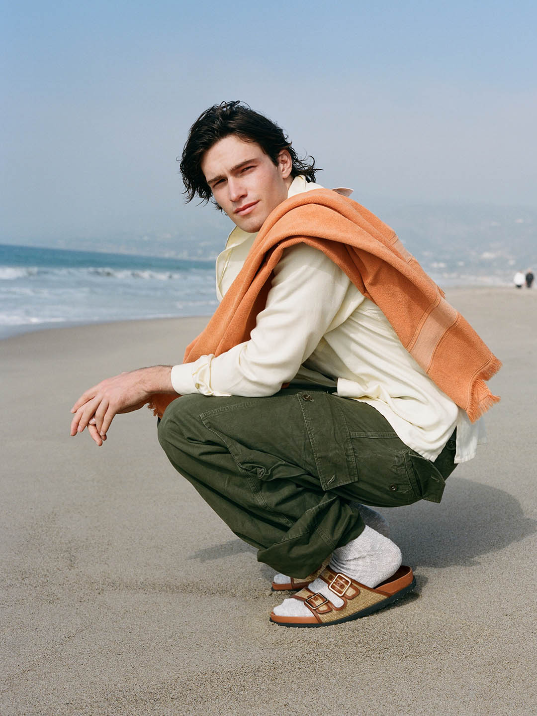 Young man squatting on a sandy beach wearing cargo trousers, a white shirt, an orange towel over his shoulders and sandals with socks