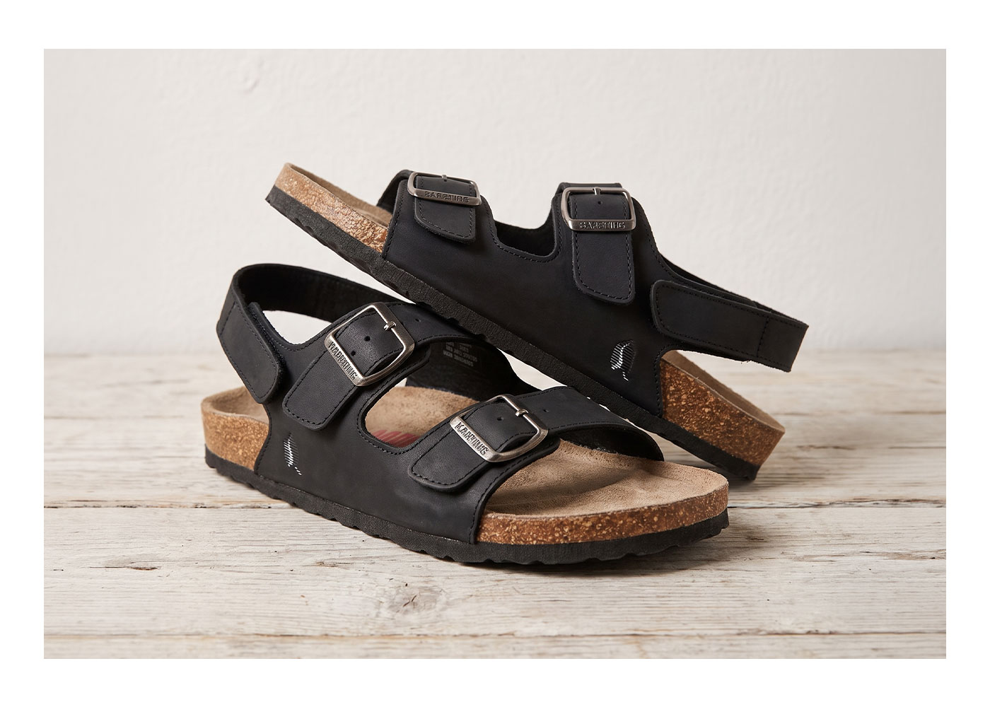 Black cork-footbed sandals with three adjustable metal buckles and suede footbed, displayed on a weathered wooden surface
