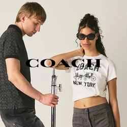 Un uomo si appoggia a un supporto per luci mentre una donna con occhiali da sole tiene una fotocamera in uno studio semplice con pareti e pavimento chiari.