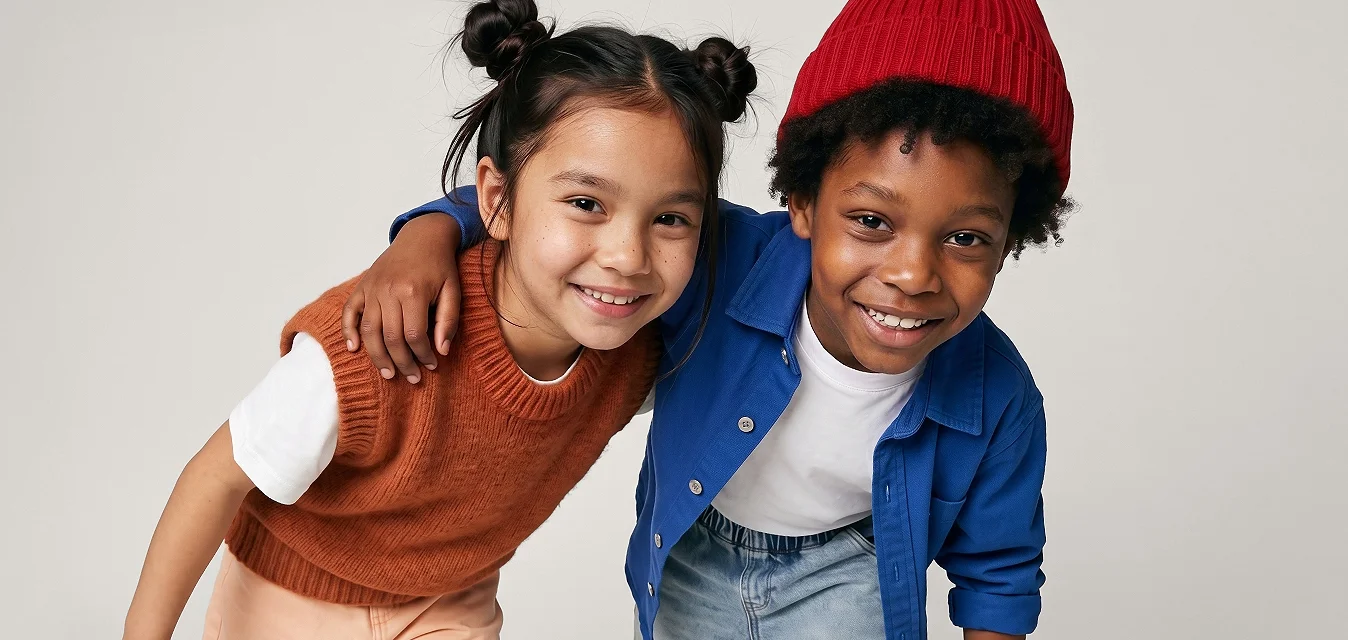 Due bambini sorridenti, uno con un berretto rosso e giacca blu, l’altro con un gilet di maglia color ruggine, piegati in avanti con le braccia intorno l’uno all’altro.
