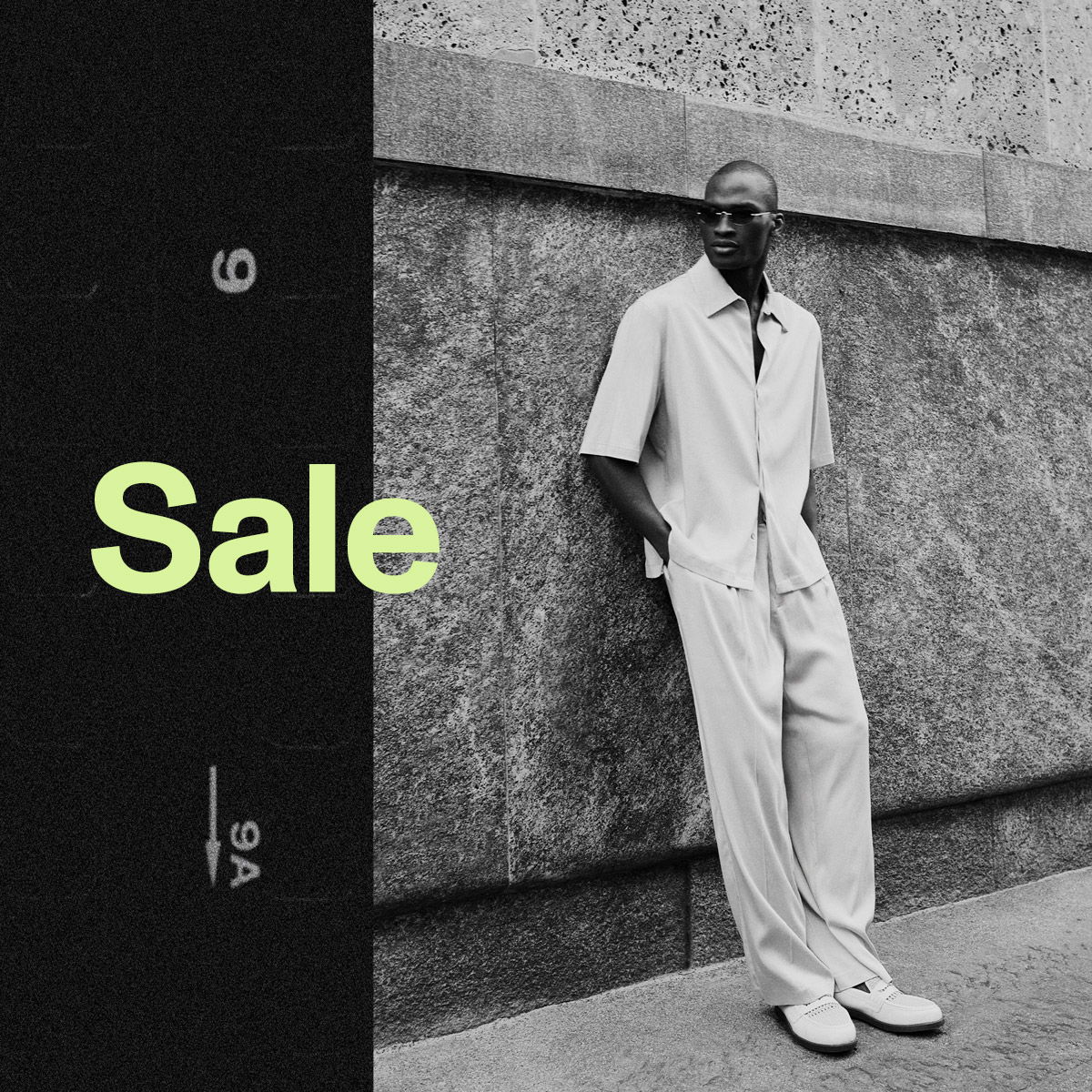 Monochrome image of a male model in a relaxed summer shirt and wide trousers leaning against a stone wall with sunglasses and 'Sale' text overlay