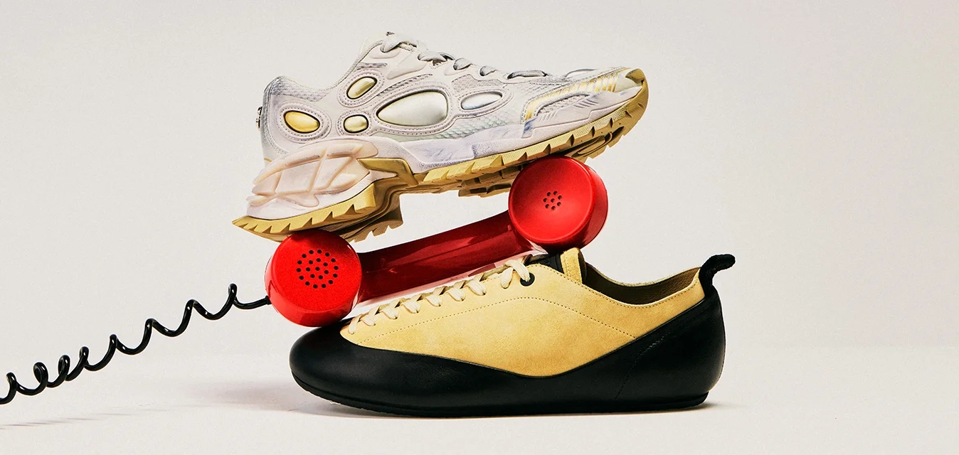 Un ricevitore telefonico vintage rosso bilanciato tra due scarpe da tennis, una beige e nera e l'altra grigio chiaro con dettagli gialli, su uno sfondo semplice.