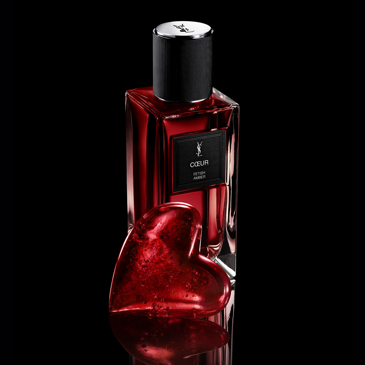 Un flacone di YSL Coeur Eau de Parfum dietro a una scultura a forma di cuore in cristallo rosso brillante su sfondo nero.