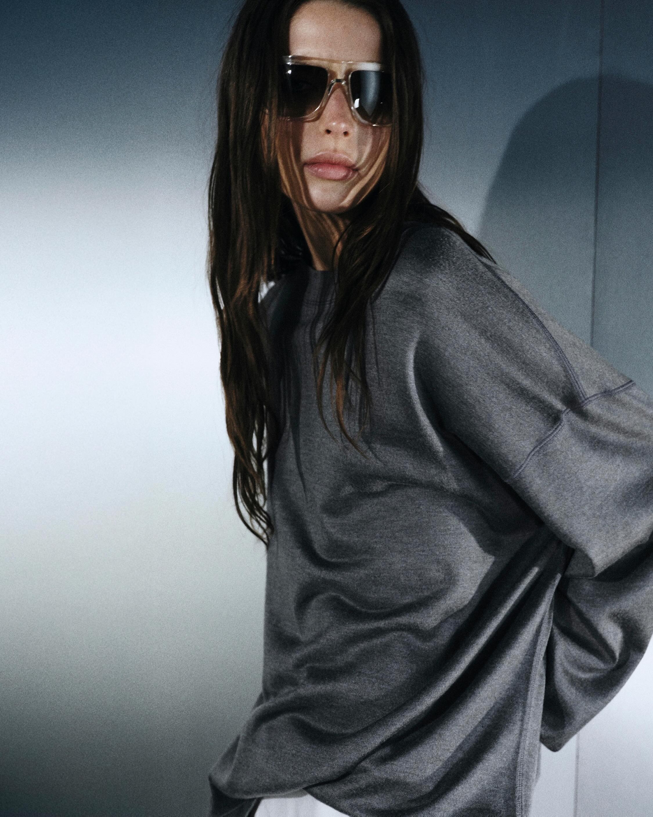 Woman in oversized grey sweater and sunglasses with long brown hair, fashion portrait against a pale wall