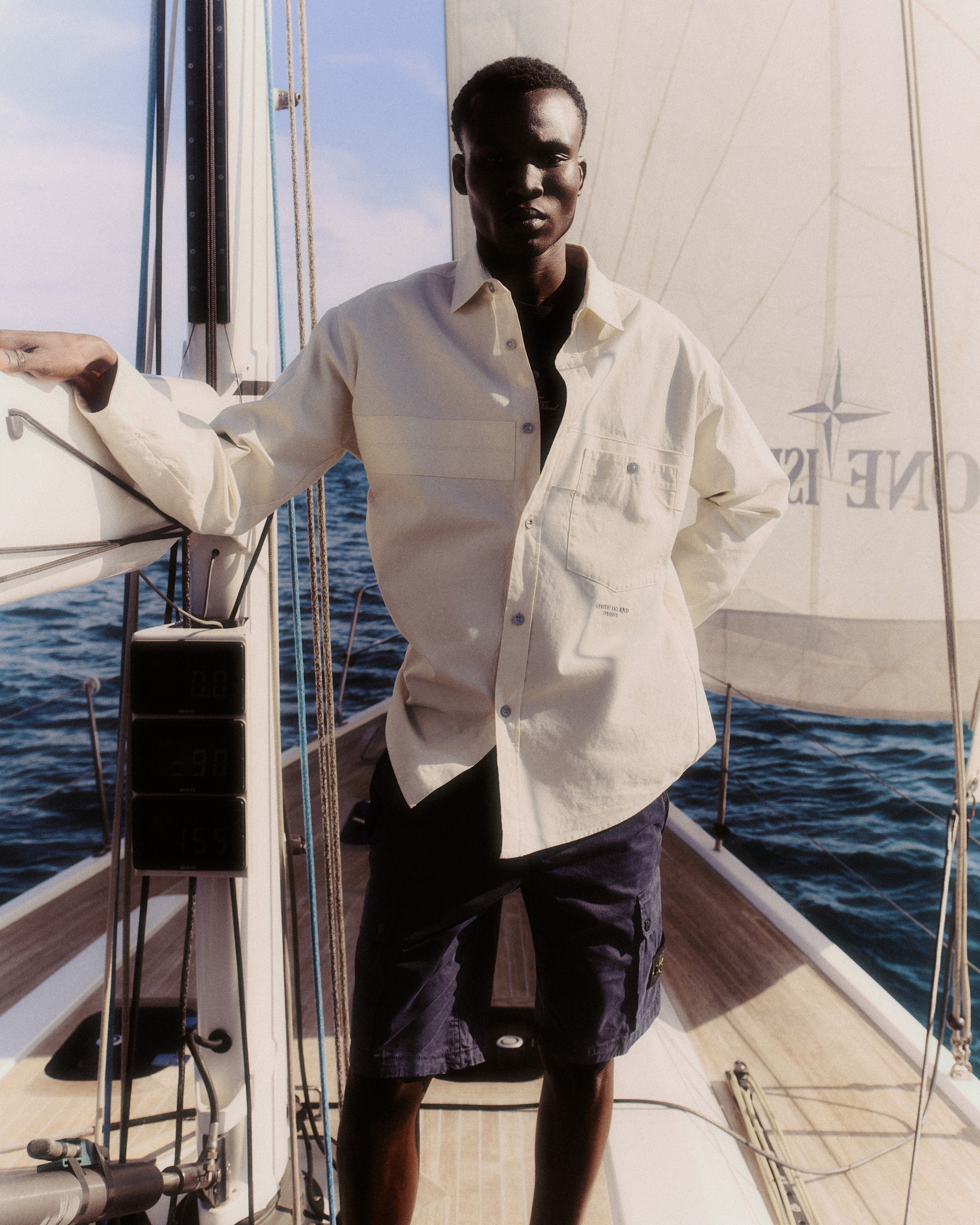 Man standing on a sailboat deck wearing a cream shirt and navy shorts, leaning on the mast with sails and open sea in the background