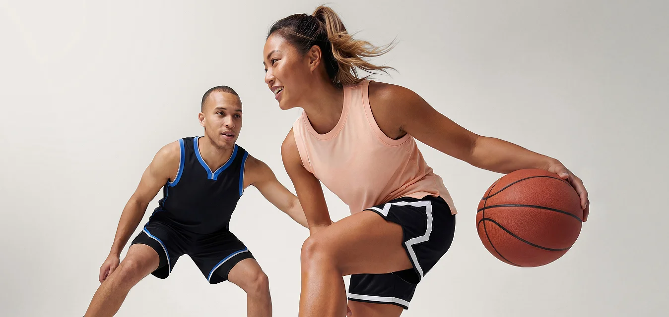 Donna che dribbla una palla da basket mentre un uomo in posizione difensiva la difende su uno sfondo chiaro e uniforme.