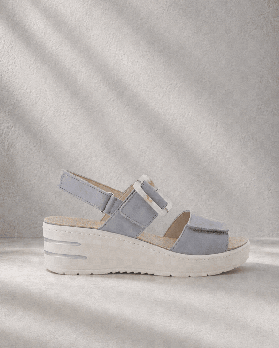Side-by-side studio photo of a light grey wedge sandal and a brown sporty open-toe sandal on textured backgrounds