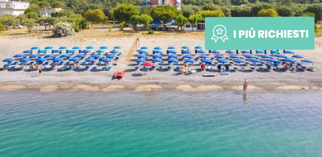 Aerial view of organised pebble beach with rows of blue umbrellas and sunbeds by turquoise sea