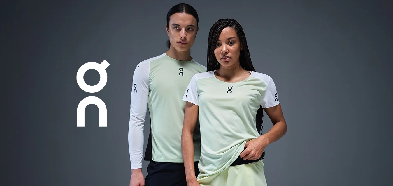 Due modelli che indossano magliette sportive verde chiaro e bianche con loghi, in piedi con sicurezza contro uno sfondo grigio scuro.