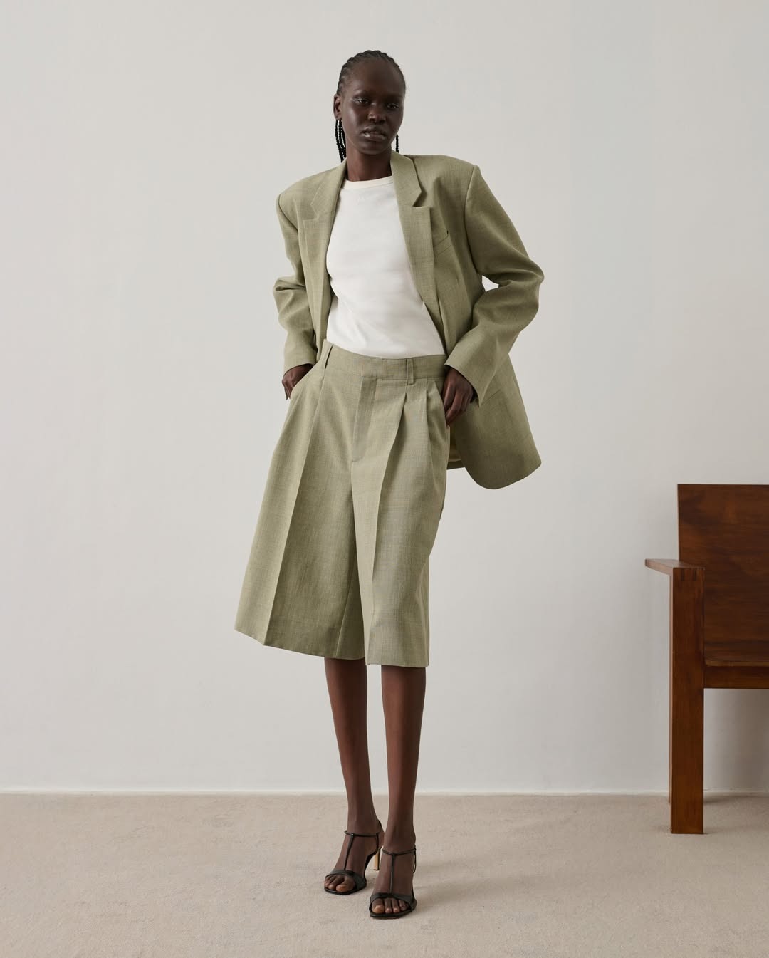 Model wearing sage-green oversized blazer and pleated shorts, white T-shirt and black strappy heels on plain background
