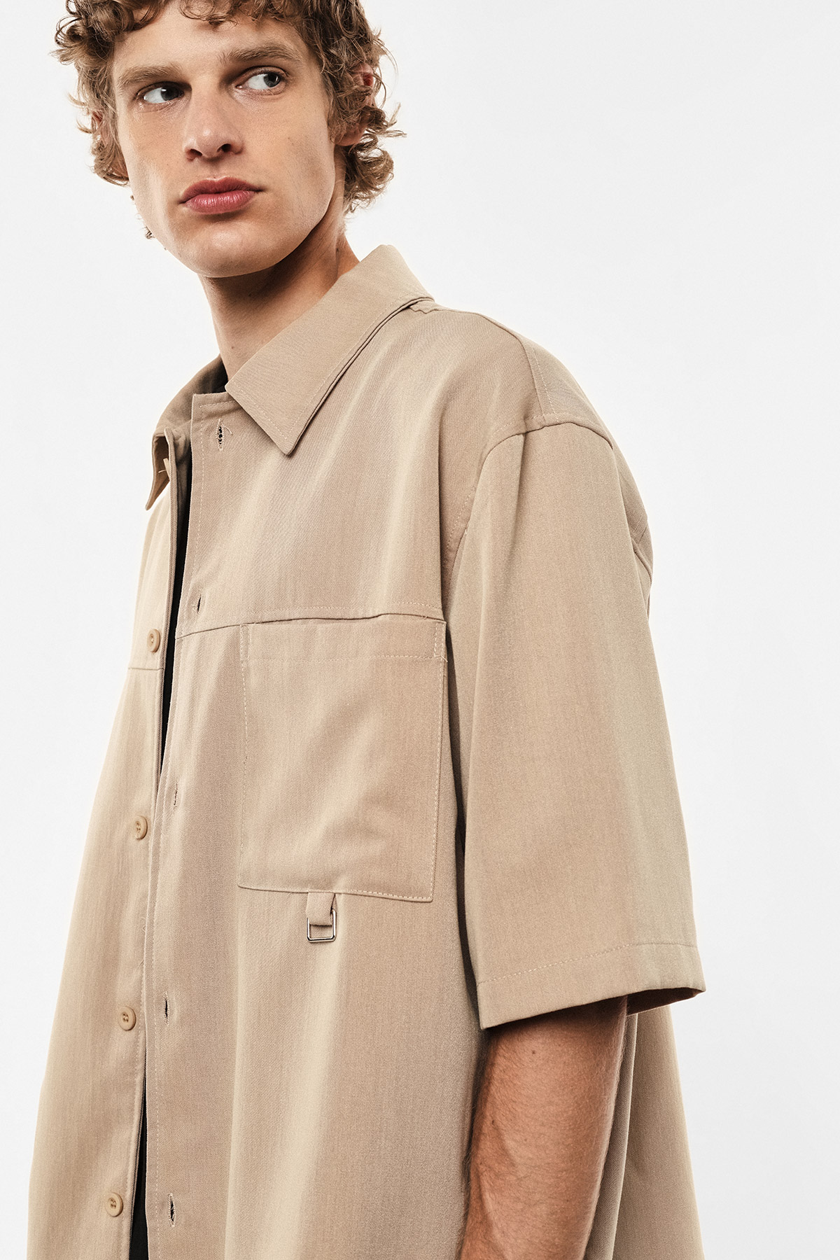 Male model wearing beige short-sleeve overshirt with chest pockets, standing against white background, looking left