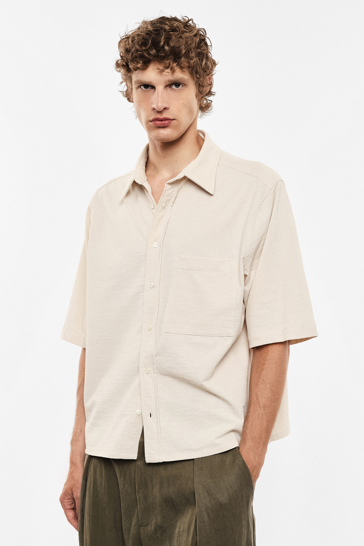 Male model wearing beige short-sleeve button-up shirt and olive trousers, hands in pockets, neutral expression on white background