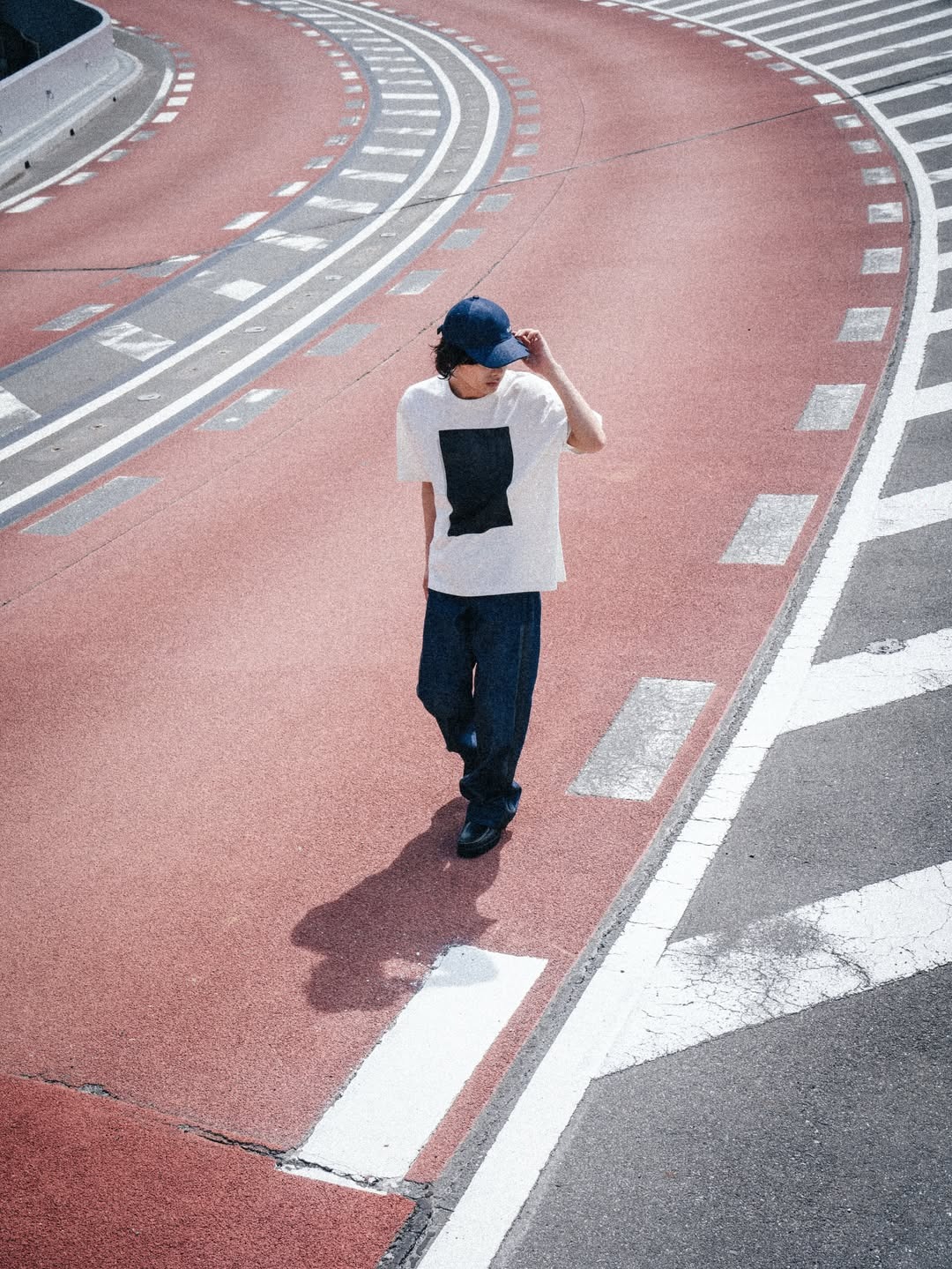 Person in a cap walking alone on a curved red urban bike lane with white road markings and long shadow