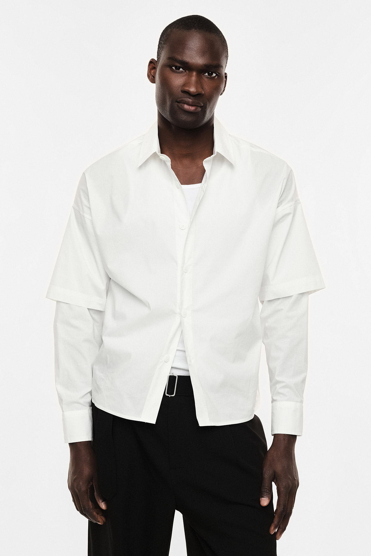 Male model wearing a white layered button-up shirt over a long-sleeve tee and black trousers, standing against a plain white background