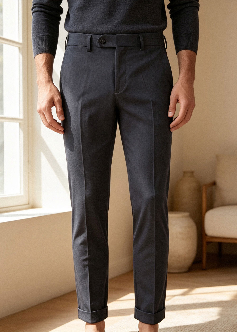 Man wearing navy tailored cuffed trousers and dark sweater, standing by sunlit window in modern room — smart menswear
