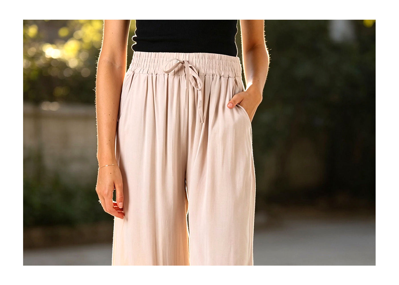 Close-up of a person wearing beige wide-leg drawstring trousers and a black top, one hand in pocket in soft outdoor light