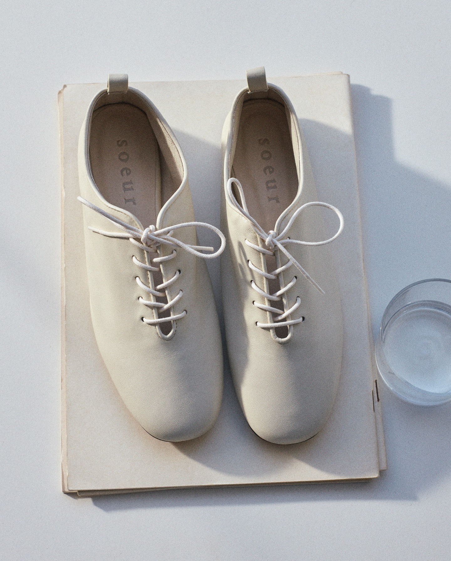 Top-down view of beige suede lace-up women's shoes on stacked cream papers beside a glass of water