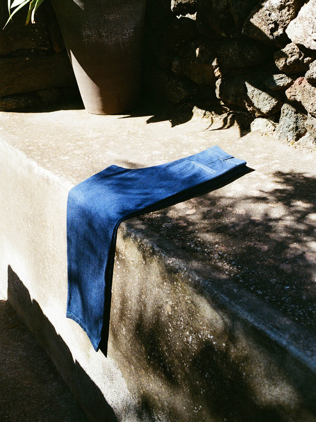 Blue denim jeans folded over a sunlit stone bench beside a clay pot and rocky wall in an outdoor garden setting