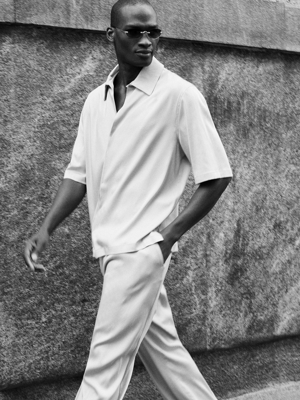 Monochrome photo of a man in sunglasses wearing a relaxed short-sleeve shirt and trousers walking past a stone wall