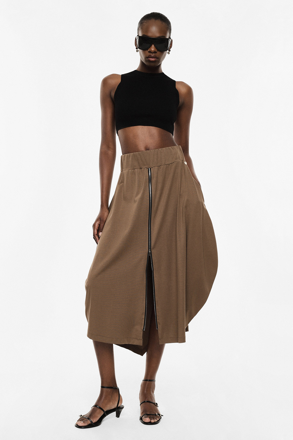 Model wearing black sleeveless crop top, brown asymmetric midi skirt with central zip and front split, black sunglasses and strappy sandals