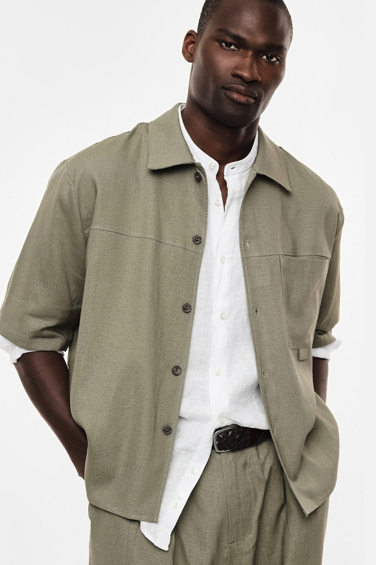 Black male model wearing sage-green relaxed overshirt over a white linen shirt and matching trousers, hands in pockets