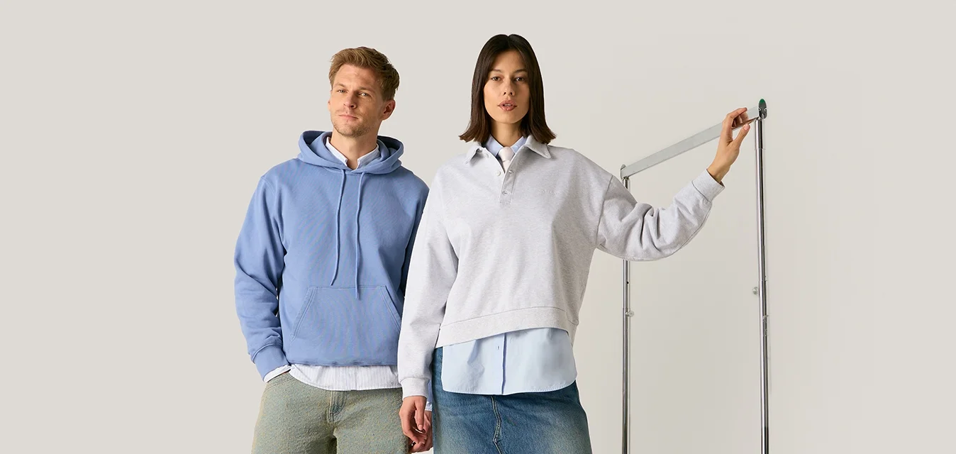 Uomo con felpa blu con cappuccio e pantaloni verdi e donna con felpa bianca e gonna di jeans in piedi, donna che tiene un dispositivo per misurare l'altezza.