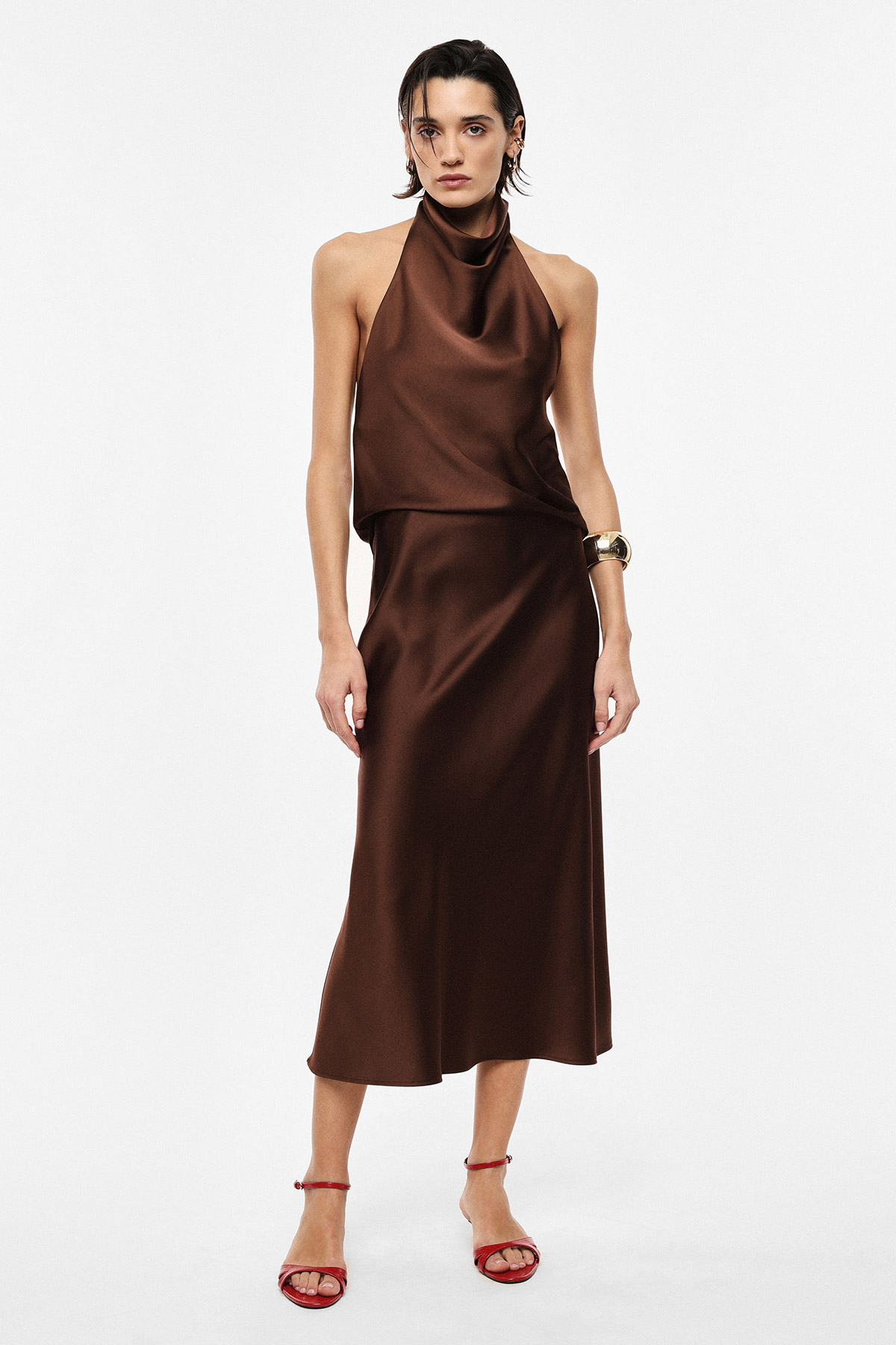 Model wearing brown satin halter-neck midi dress with red strappy heels and gold cuff bracelet on white background