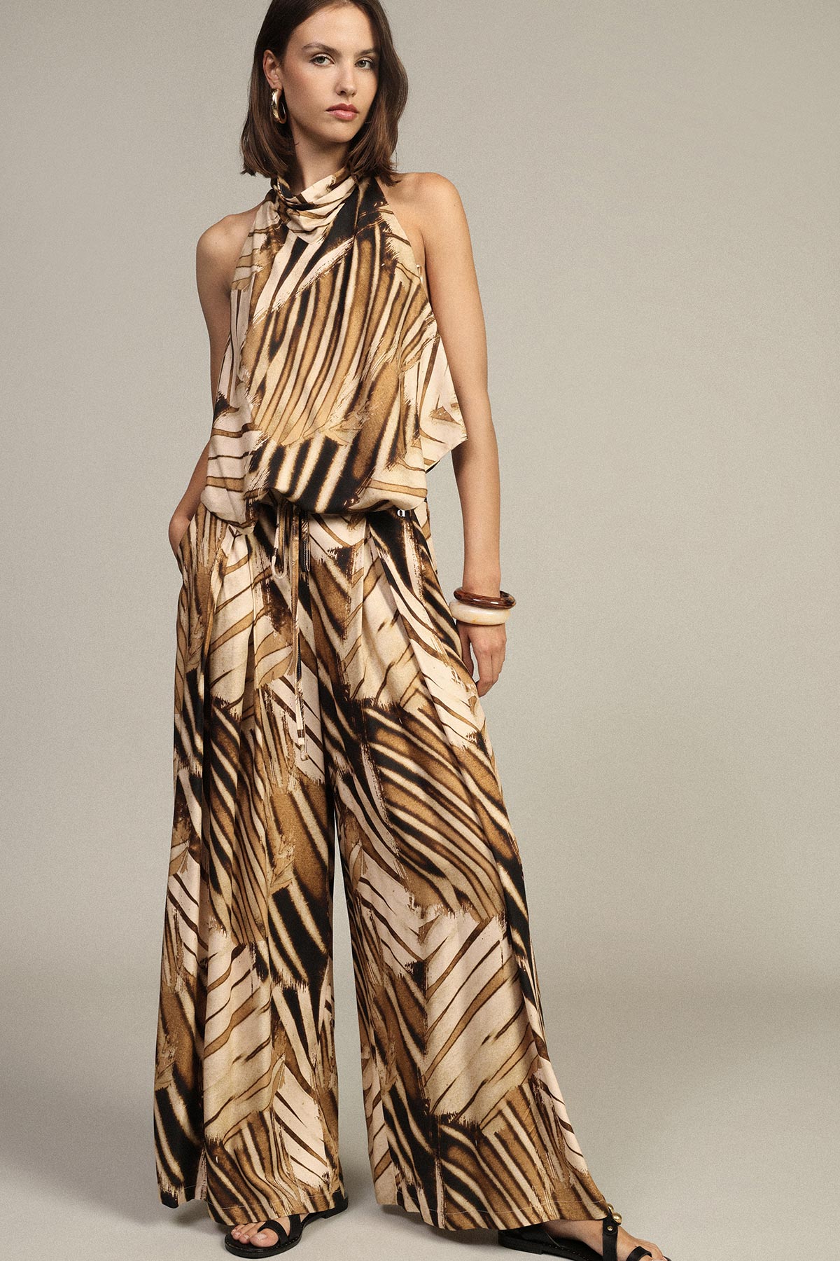 Woman wearing brown and beige abstract palm-print halter top and wide-leg trousers, hands in pockets, styled with bangles