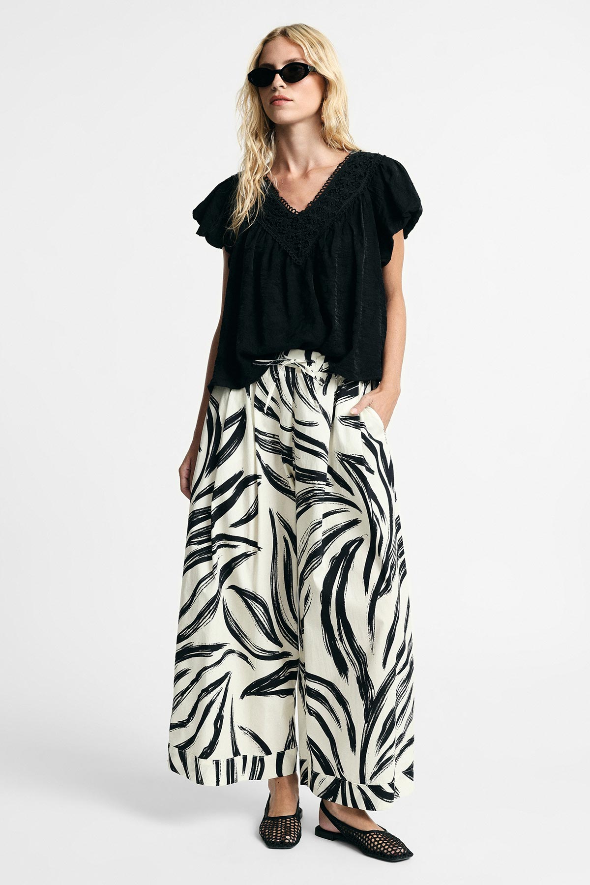 Blonde woman in black V-neck blouse and wide-leg cream trousers with black leaf print, wearing sunglasses and woven black flats