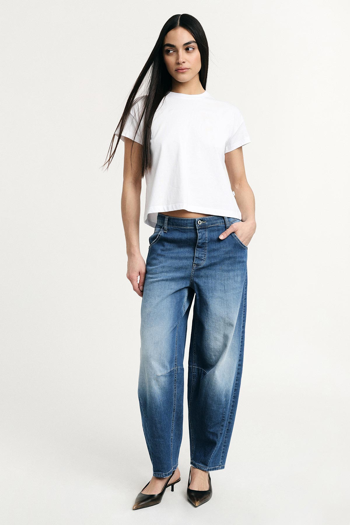 Woman in white cropped t-shirt and relaxed-fit blue jeans with black heels, standing against plain light background